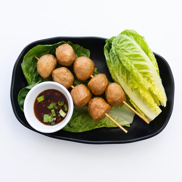Fried fish balls on skewers with lettuce and a dipping sauce, served on a black plate.