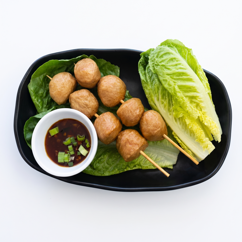 Fried fish balls on skewers with lettuce and a dipping sauce, served on a black plate.