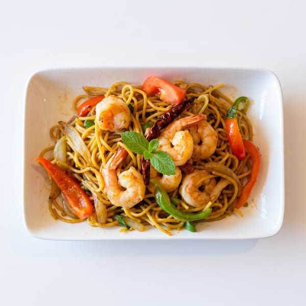 Shrimp lo mein with noodles, bell peppers, onions, and mint on a white rectangular plate.
