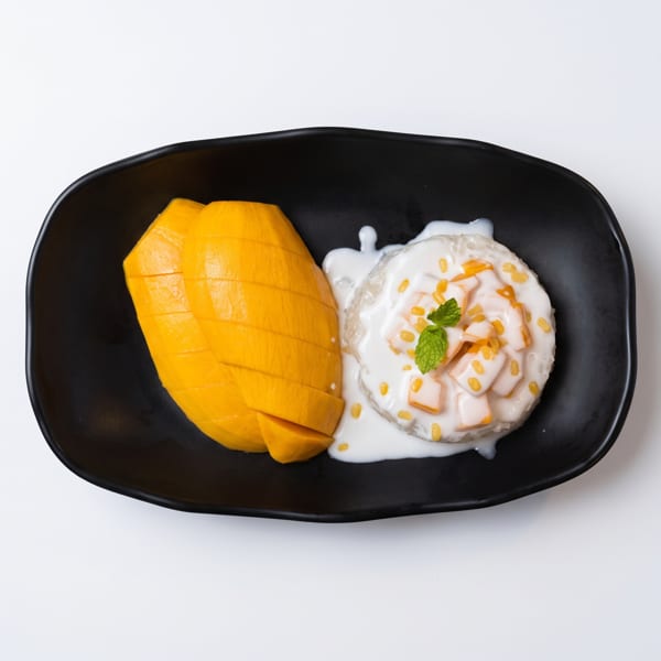 Mango sticky rice with coconut milk, topped with mung beans and a mint leaf, on a black plate.