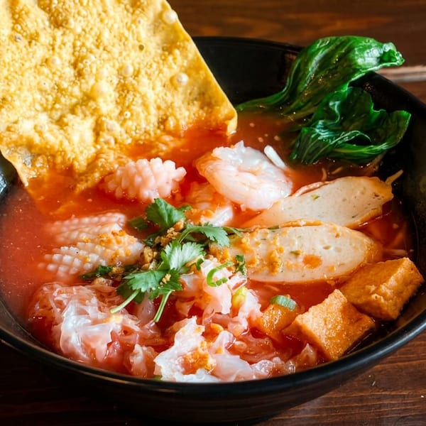 A vibrant bowl of Yen Ta Fo noodle soup with seafood, tofu, and greens on a wooden table.