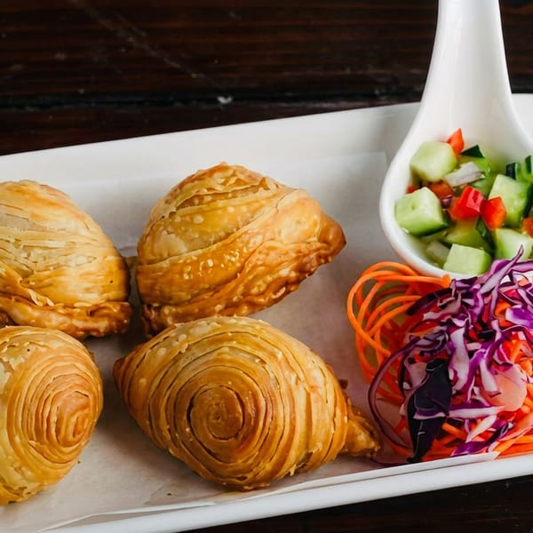 Four golden-brown curry puffs with flaky pastry, served with a side of cucumber salad and shredded vegetables.