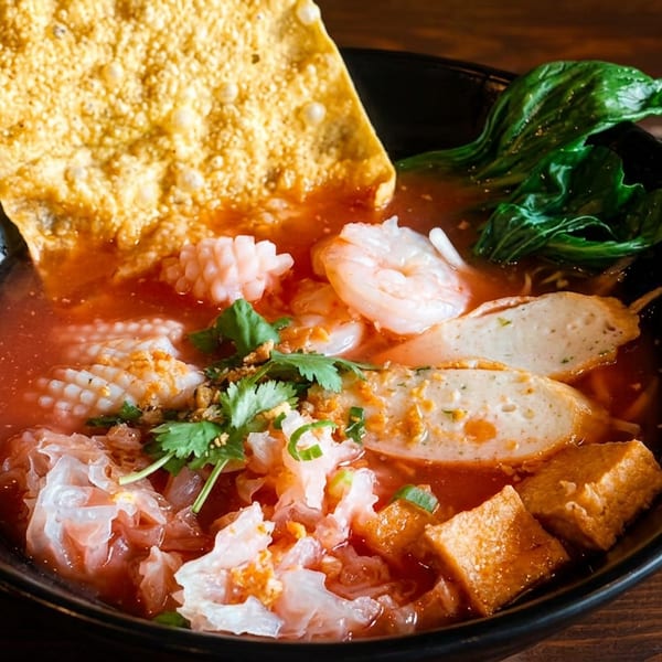 A vibrant red soup with shrimp, squid, fish cakes, tofu, and greens, garnished with a crispy wonton.
